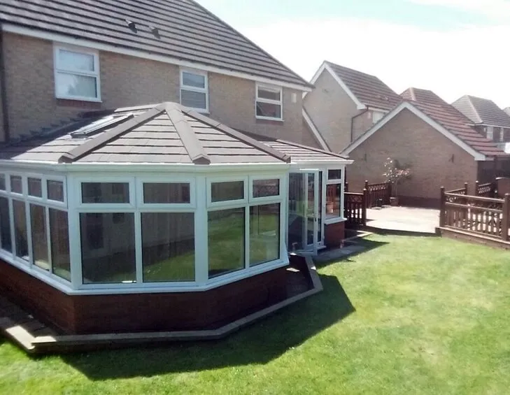 A large white conservatory with a dark tiled roof and a roof window, set on a brick base beside a lawn at a house in Benton.