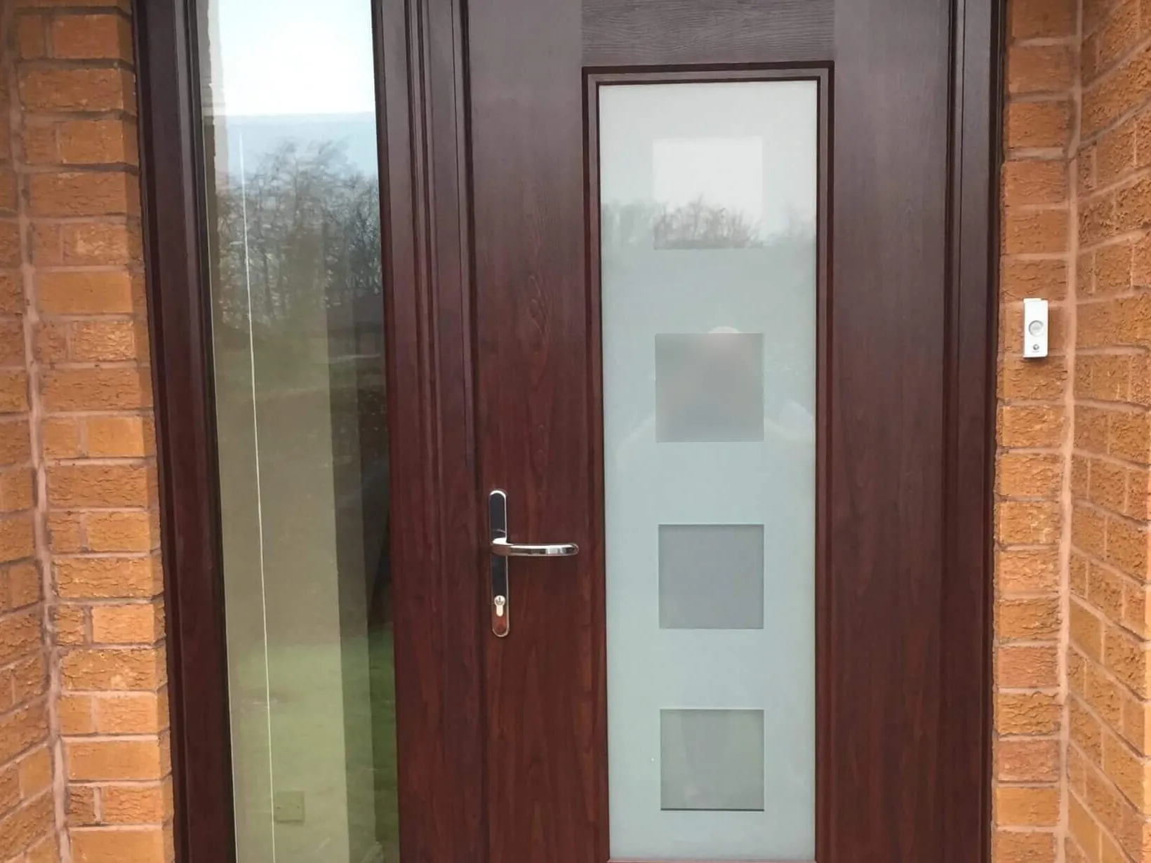 A dark wood-effect front door with a tall frosted glazed panel and chrome lever handle, beside a full-height glazed side panel, in a brick entrance in Carlisle.