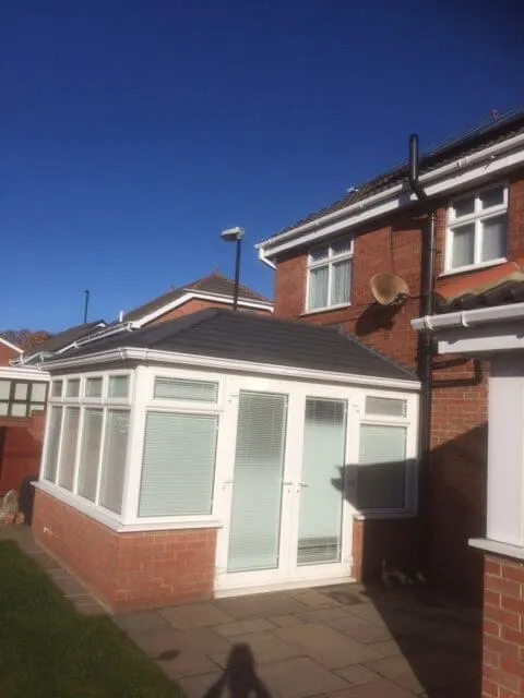A small white conservatory with a dark grey tiled roof and glazed double doors, built onto a brick house in Corbridge under a clear sky.