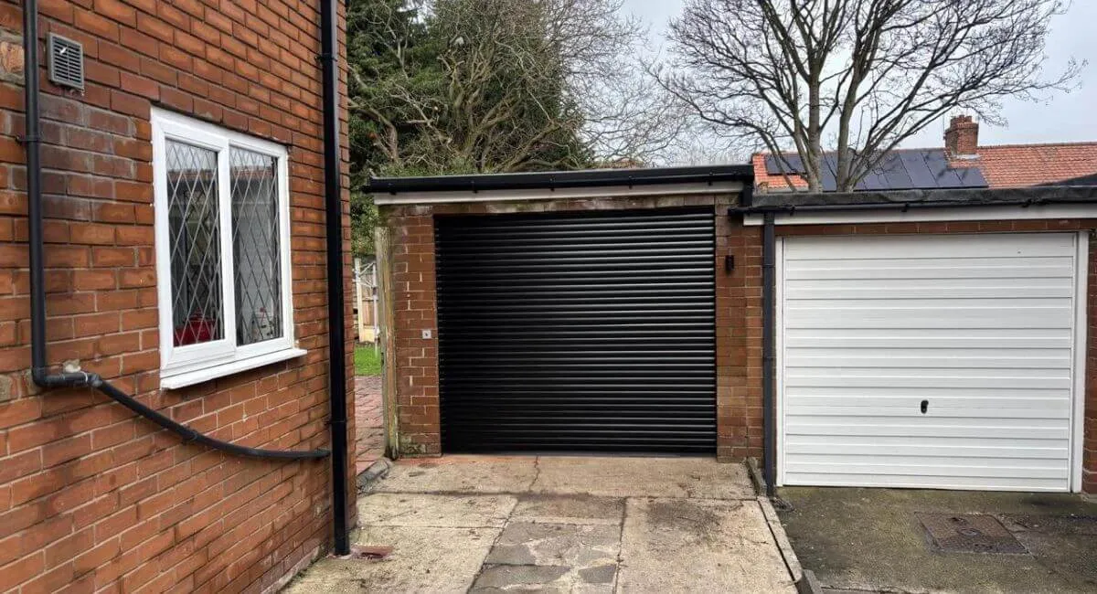 A brick garage in Gosforth, Newcastle upon Tyne with a new black roller-style door and a finished flat-roof edge with guttering, seen from the driveway on an overcast day.