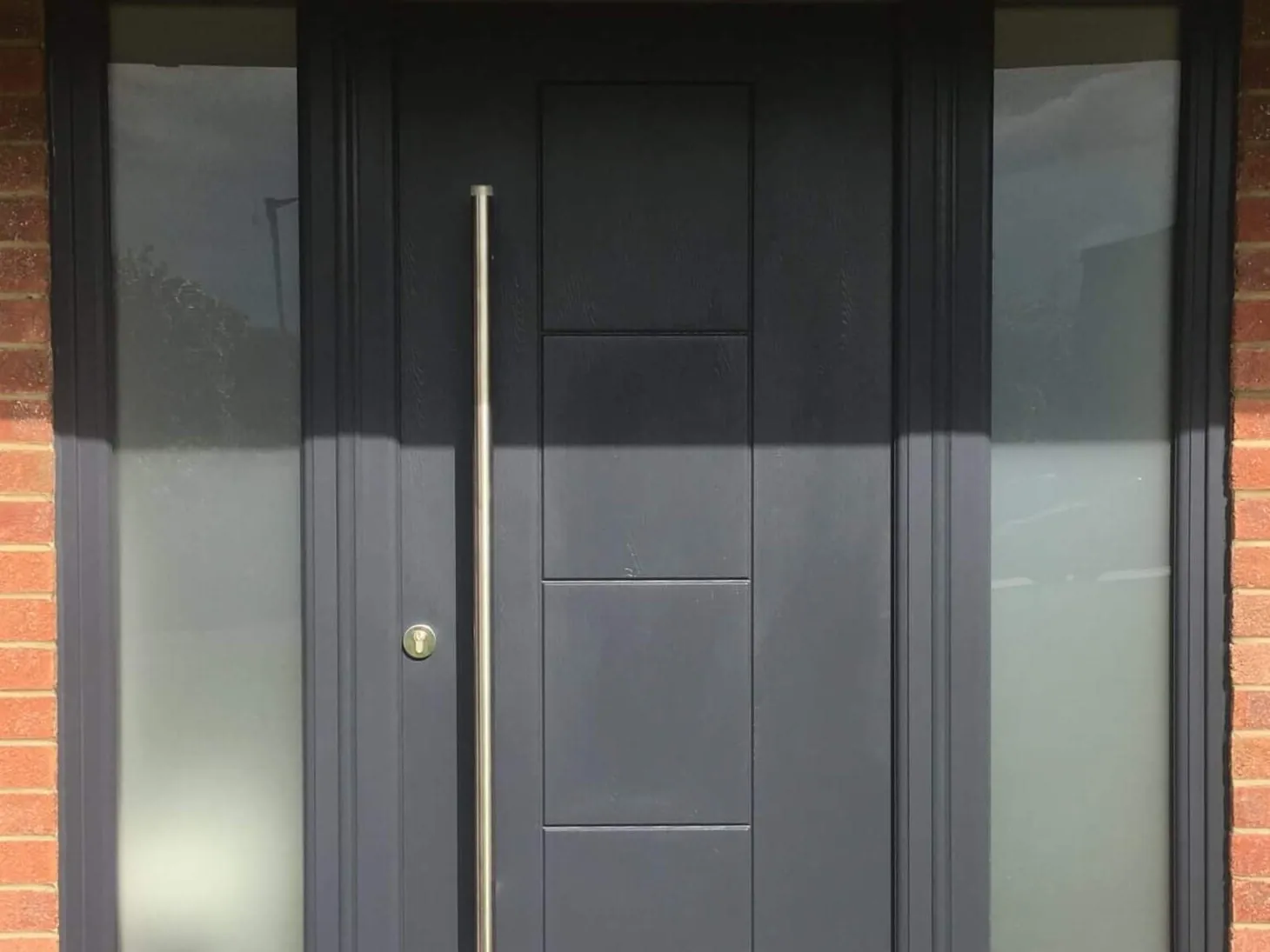 A dark grey flush front door with a full-height bar handle, flanked by two tall glazed side panels in a matching dark frame, in a red-brick elevation in Seahouses.