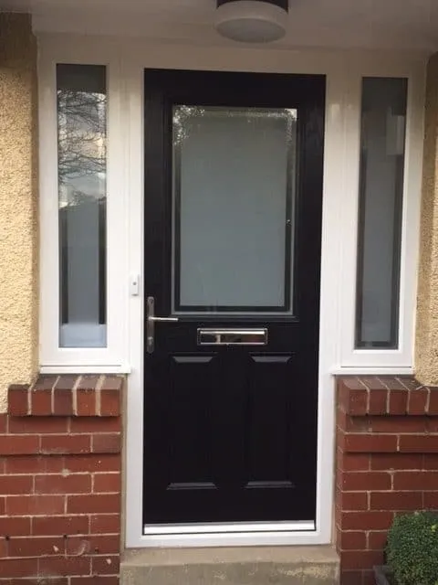 A dark front door with an obscured glazed panel and chrome furniture in Tynemouth, set in a white frame between two tall glazed sidelights on low brick plinths.